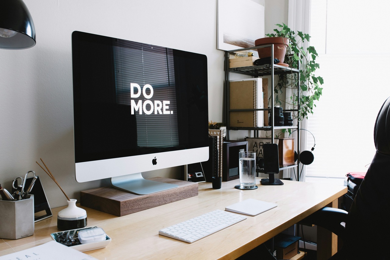 A sleek desk with a modern iMac displaying "DO MORE." surrounded by plants, stationery, and a glass of water.